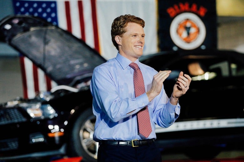 Rep. Joe Kennedy III claps on stage in front of a car and an American flag.